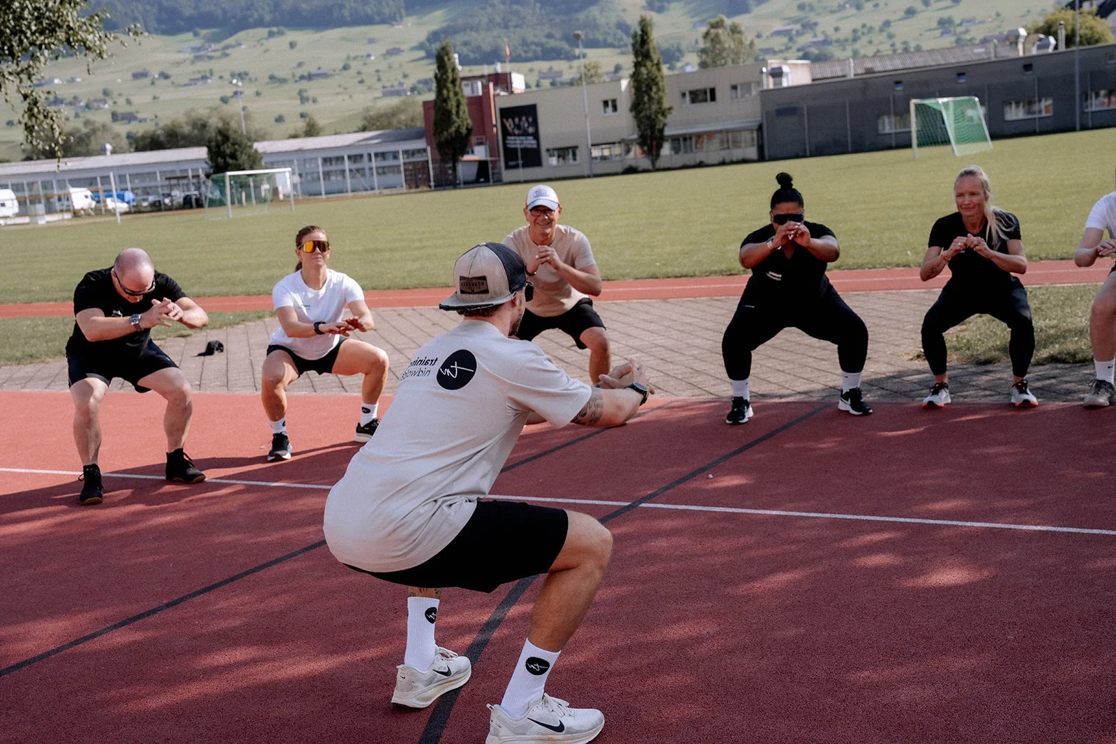 A coach in a training nidwalden shirt demonstrating a squat on the running track while the group follows along